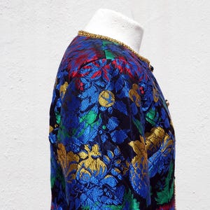 Pode incluir: Casaco vintage com um padr&atilde;o floral vibrante em azul, dourado, vermelho e verde. O casaco tem uma borda dourada ao redor da gola e &eacute; exibido em um manequim. O tecido tem uma apar&ecirc;ncia texturizada.