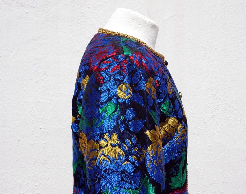 Pode incluir: Casaco vintage com um padr&atilde;o floral vibrante em azul, dourado, vermelho e verde. O casaco tem uma borda dourada ao redor da gola e &eacute; exibido em um manequim. O tecido tem uma apar&ecirc;ncia texturizada.