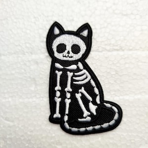 Op de afbeelding: Zwart-wit geborduurde patch met een katten-skelet ontwerp. De patch toont een zittende kat met een gedetailleerde skeletstructuur, wit omlijnd tegen een zwarte achtergrond. De patch heeft een zwarte rand.