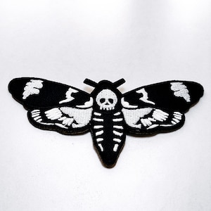 May include: Black and white embroidered patch in the shape of a moth. The moth design features a skull and skeleton details on the body and wings. The patch is likely for clothing or accessories.