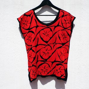 May include: A sleeveless red top with black floral embroidery. The top is hanging on a black hanger against a white wall. The embroidery features a repeating pattern of stylized flowers.