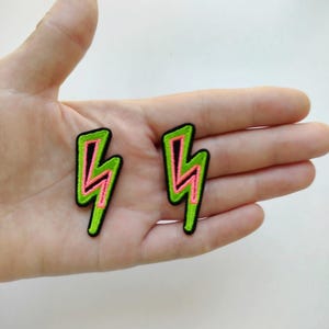 Puede incluir: Dos parches bordados con forma de relámpagos. Cada parche tiene un contorno negro, un borde verde brillante y un diseño de rayo rosa en el centro. Los parches se muestran sobre un fondo claro.