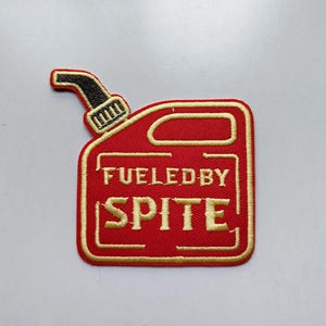 May include: Embroidered patch in the shape of a red gas can with a black spout. The text "FUELED BY SPITE" is embroidered in gold thread. The patch has a gold border and is on a white background.