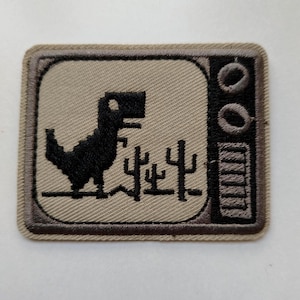 Puede incluir: Parche bordado con un dinosaurio negro pixelado sobre un fondo beige, enmarcado por un borde gris oscuro. El diseño incluye caracteres chinos y un marco tipo TV, con un indicador de batería en el lado derecho.