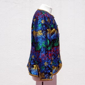 Pode incluir: Casaco floral vibrante em tons de azul, verde, vermelho e dourado. O casaco apresenta decote redondo e acabamentos dourados. O tecido tem um aspeto texturizado e o casaco &eacute; abotoado.