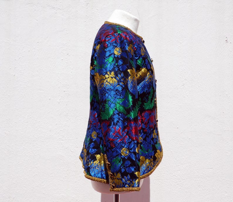Pode incluir: Casaco floral vibrante em tons de azul, verde, vermelho e dourado. O casaco apresenta decote redondo e acabamentos dourados. O tecido tem um aspeto texturizado e o casaco &eacute; abotoado.