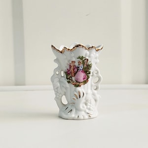 Pode incluir: Um pequeno vaso de porcelana branca com borda recortada e acabamento dourado. O corpo é decorado com uma impressão colorida de duas figuras num jardim, acentuado por motivos em relevo de uvas e folhas com detalhes pintados a ouro.