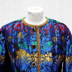 Pode incluir: Casaco vintage com um vibrante padr&atilde;o floral em azul, vermelho, verde e dourado. O casaco apresenta decote redondo, bot&otilde;es pretos e acabamento dourado. O tecido tem uma textura acolchoada.