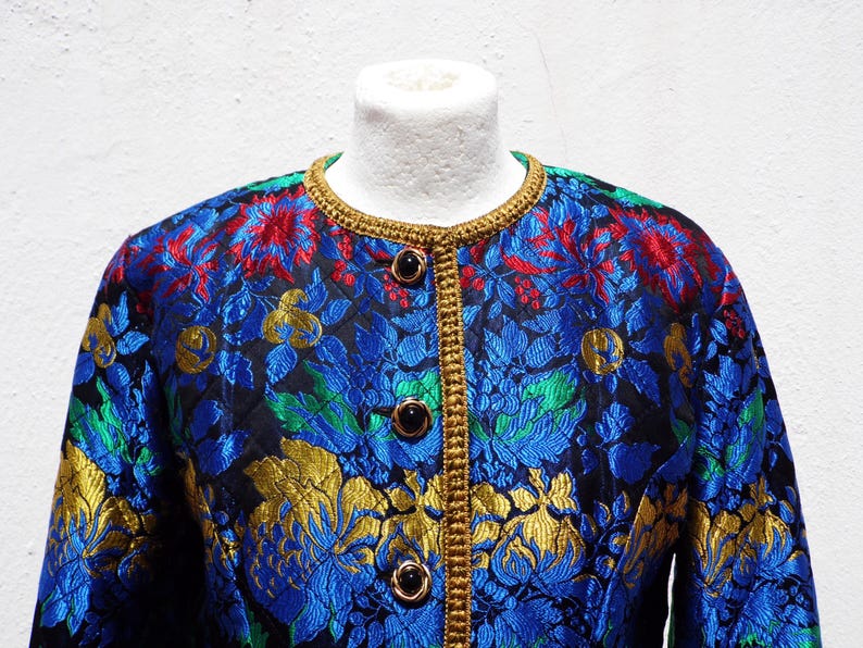 Pode incluir: Casaco vintage com um vibrante padr&atilde;o floral em azul, vermelho, verde e dourado. O casaco apresenta decote redondo, bot&otilde;es pretos e acabamento dourado. O tecido tem uma textura acolchoada.