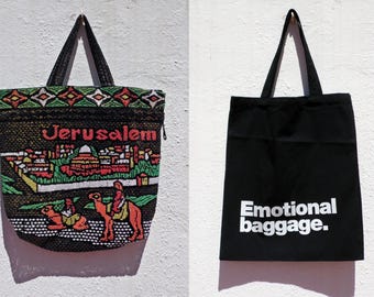 May include: Two tote bags hanging against a white wall. One is black with the text "Emotional baggage." in white. The other is a beaded bag with the word "Jerusalem" and an image of a city and camels.