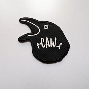 May include: Black crow-shaped patch with white embroidered details. The crow's beak is open, and the word "CAW-f" is embroidered in white. The patch is likely for clothing or accessories.