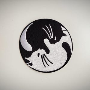 Può includere: Toppa ricamata con un design yin-yang di due gatti. Un gatto è bianco, l'altro nero, disposti in un motivo circolare. La toppa ha uno sfondo nero e un bordo bianco.