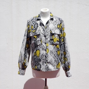 Op de afbeelding: Een overhemd met lange mouwen en knoopsluiting, voorzien van een slangenprint in grijze, zwarte en felgele tinten. Het overhemd heeft een klassieke kraag, twee borstzakken met klep en knoopmanchetten, getoond op een paspop.