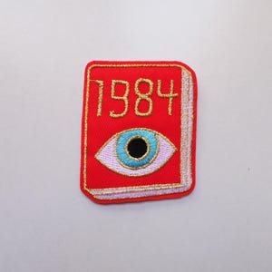 May include: A red embroidered patch with the gold text "1984" above a stylized eye with a blue iris and black pupil. The patch is book-shaped with gold and pink trim. Ideal for customising clothing or accessories.