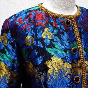 Pode incluir: Um close-up de um casaco com padr&atilde;o floral. O casaco apresenta um fundo preto com desenhos florais azuis, vermelhos, verdes e dourados. Possui uma guarni&ccedil;&atilde;o dourada ao redor da gola e na frente, com bot&otilde;es pretos.