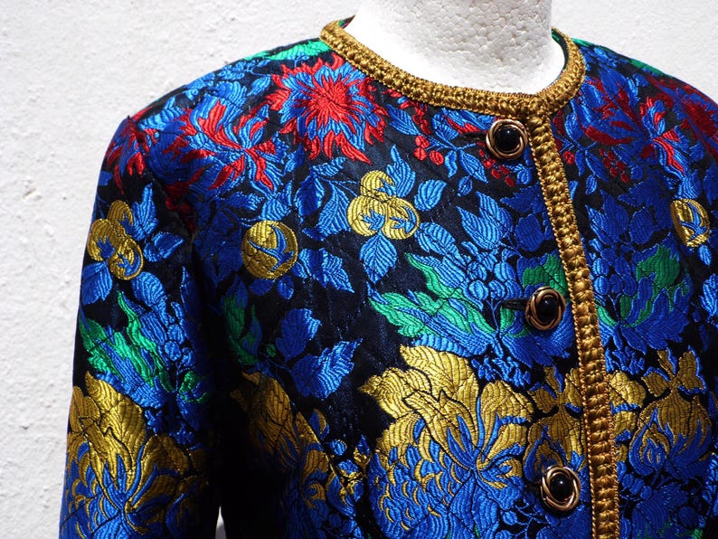 Pode incluir: Um close-up de um casaco com padr&atilde;o floral. O casaco apresenta um fundo preto com desenhos florais azuis, vermelhos, verdes e dourados. Possui uma guarni&ccedil;&atilde;o dourada ao redor da gola e na frente, com bot&otilde;es pretos.
