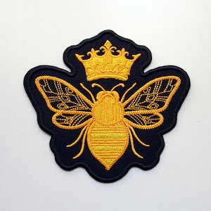 May include: Embroidered patch of a golden bee wearing a crown, on a navy blue background. The bee's wings and body are detailed with yellow stitching. The patch has a unique, stylized shape.
