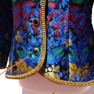 Pode incluir: Um close-up de um casaco vintage com um padr&atilde;o floral em azul, vermelho, verde e dourado sobre um fundo preto. O casaco tem acabamentos dourados e bot&otilde;es pretos.