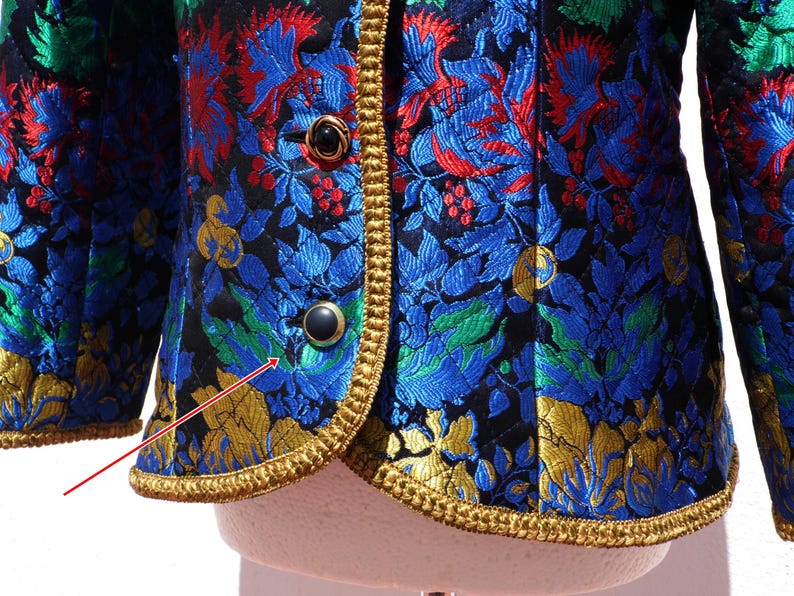 Pode incluir: Um close-up de um casaco vintage com um padr&atilde;o floral em azul, vermelho, verde e dourado sobre um fundo preto. O casaco tem acabamentos dourados e bot&otilde;es pretos.
