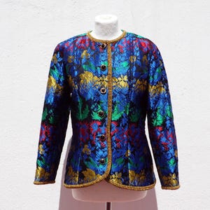 Pode incluir: Casaco vintage com padr&atilde;o floral em tons de azul, verde, vermelho e dourado. O casaco apresenta decote redondo, acabamento dourado e bot&otilde;es decorativos. A pe&ccedil;a &eacute; exibida em um manequim sobre um fundo branco.