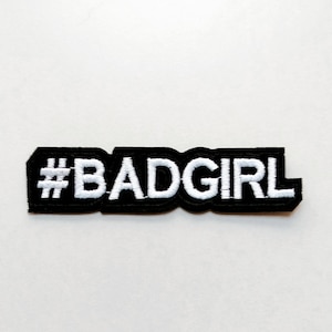Puede incluir: Un parche negro con el texto "#BADGIRL" en blanco, cosido en el parche. El parche es rectangular con esquinas redondeadas. El fondo es blanco liso.
