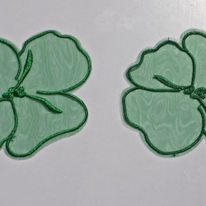 Op de afbeelding: Twee groene bloemvormige applicaties met glinsterende groene contouren en details. De applicaties hebben een doorschijnend, getextureerd uiterlijk en zijn geplaatst tegen een witte achtergrond. Het bloemmotief heeft bloemblaadjes en een centrale stengel.