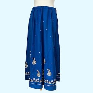 May include: A long, flowing blue skirt with white polka dots and an embroidered paisley design along the hemline. The skirt has a drawstring waist and a gold trim. The skirt is made of a lightweight fabric.