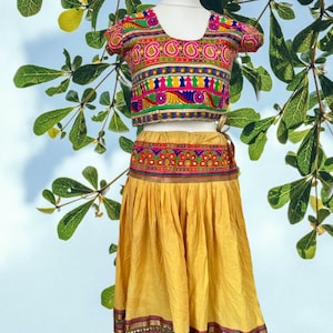 May include: A two-piece outfit consisting of a short-sleeved crop top and a long, pleated skirt. The top features vibrant, multi-colored embroidery in pink, yellow, blue, and green. The skirt is solid mustard yellow with a matching embroidered hem.
