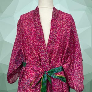 Può includere: Un kimono color magenta caratterizzato da un fitto e intricato motivo floreale nei toni del rosa, giallo e blu. Il capo presenta maniche ampie, uno scollo a V e una cintura in tessuto verde a contrasto annodata in vita con un bordo dorato.