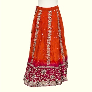 May include: An orange and fuchsia skirt with intricate embroidery and embellishments. The skirt features vertical embroidered patterns with silver accents and small, reflective embellishments. The bottom hem is a deep fuchsia with detailed embroidery.