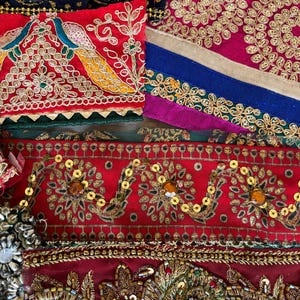 Könnte beinhalten: Eine Sammlung geschichteter Stoffborten mit aufwendigen Stickereien, goldenem Metallic-Garn, Pailletten und Perlen. Die Textilien zeigen leuchtende Farben wie Rot, Dunkelrosa, Marineblau und Grün mit Pfauen- und Blumenmotiven.