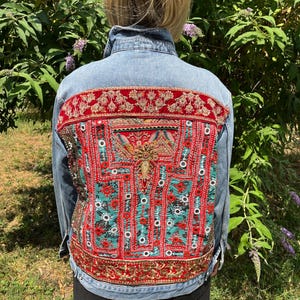 Peut inclure: Une veste en jean bleu clair dotée d'un grand panneau arrière rectangulaire orné de broderies complexes en tissu rouge et bleu sarcelle, de détails en fil doré métallique, de petits miroirs circulaires et de motifs floraux sur les bordures.