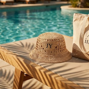 May include: A woven straw bucket hat with the initials "JY" in black is displayed on a wooden surface. A canvas tote bag is next to the hat. The background features a swimming pool with clear blue water.