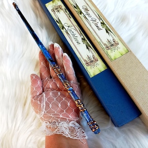 Op de afbeelding: Een blauwe en gouden toverstaf met een wervelend ontwerp, vastgehouden in de buurt van een blauwe doos met de woorden "Magic Wand" en "Kathleen" erop gedrukt. De toverstaf heeft koperkleurige accenten en is ongeveer 30 cm lang.