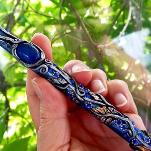 Puede incluir: Una varita azul hecha a mano con detalles plateados y dorados, adornada con piedras preciosas azules y una gran piedra azul ovalada. La varita presenta detalles intrincados y se sostiene sobre un fondo verde borroso.