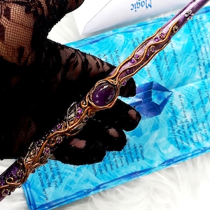 May include: A handcrafted purple and gold magic wand featuring an oval amethyst stone and decorative detailing. The wand is set against a blue backdrop with the words "Magic Letter".