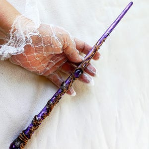 Puede incluir: Una varita mágica morada con detalles dorados y una gran gema morada. La varita presenta intrincados patrones y adornos en forma de hojas. El fondo es blanco.