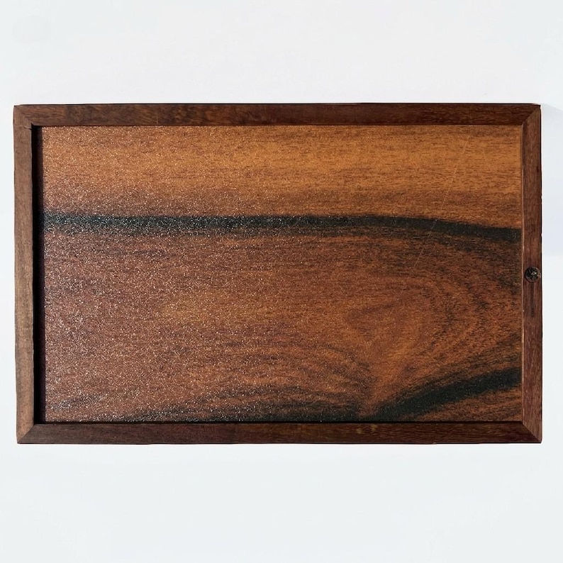 May include: A rectangular wooden tray featuring a rich, warm brown grain pattern with prominent dark, nearly black horizontal streaks running across the center. The tray is enclosed by a solid, dark brown wooden frame with a small metal hinge.