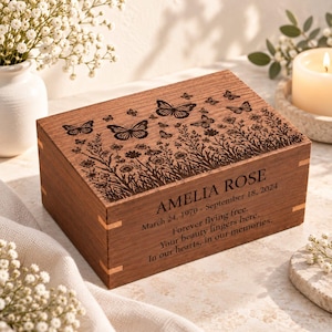 Op de afbeelding: Een rechthoekige houten aandenkendoos met een lasergegraveerd ontwerp van vlinders en wilde bloemen op het deksel. Het voorpaneel is gegraveerd met de naam AMELIA ROSE, data en de tekst Forever flying free. Your beauty lingers here.