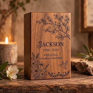 May include: A rectangular wooden cremation urn featuring laser-engraved floral branches with two birds, the name JACKSON, the years 1958-2023, the text A life of love, a memory cherished, a flying dove with a heart, and a cluster of wildflowers.