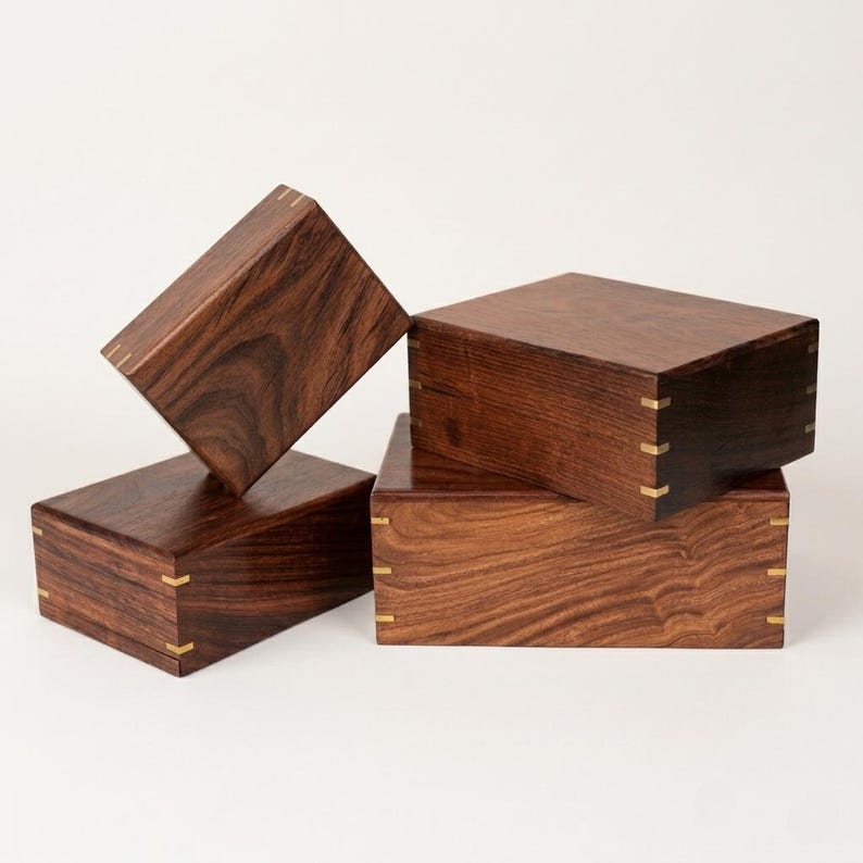 May include: Three rectangular dark brown wooden boxes with visible wood grain patterns. Each box features light-colored metal corner accents at the joints, providing a contrast to the dark wood. The boxes are arranged in a stacked and angled formation.