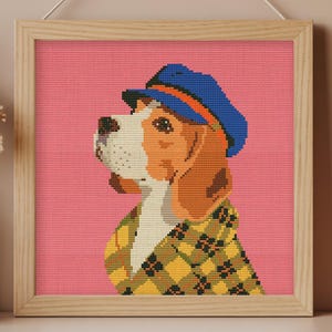 May include: A framed cross-stitch picture of a beagle wearing a blue cap and a yellow and black plaid jacket. The dog is set against a pink background. The frame is light wood. The artwork is hanging on a wall.
