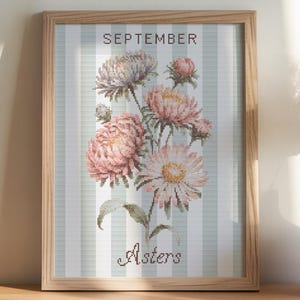 May include: A framed floral print featuring pink and white asters, with the text "September" and "Asters." The artwork is set against a light blue and white striped background, in a wooden frame. A coffee cup, notebook, and vase are also visible.