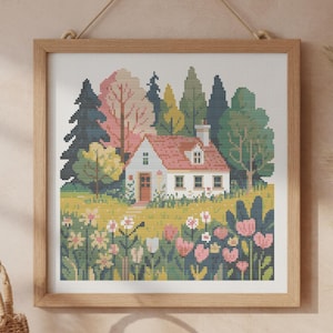 Op de afbeelding: Een ingelijste kruissteek van een huisjesscène. Het kunstwerk toont een wit huis met een rood dak, omringd door kleurrijke bloemen en bomen. De lijst is gemaakt van lichtgekleurd hout en het kunstwerk hangt aan een beige muur.