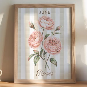 May include: A framed print featuring a cross-stitch design of pink roses with green leaves against a striped background. The word "June" is at the top, and "Roses" is at the bottom. A coffee cup, notebook, and vase are also visible.