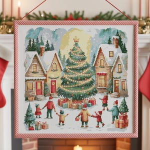 May include: A framed cross-stitch picture shows a Christmas scene with a snowy village, a decorated tree, and children playing. The frame has a red and white striped border. Red stockings hang from the mantelpiece, and a small Christmas tree is on the right.