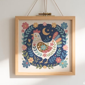 May include: A framed cross-stitch artwork depicting a hen surrounded by a floral wreath. The design includes a dark blue background with a crescent moon and stars. The artwork is in a wooden frame and is suspended from a wooden dowel.