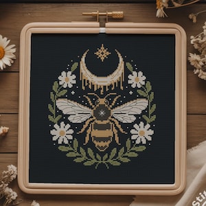 May include: Cross-stitch embroidery of a bee with a crescent moon and floral elements, set in a wooden hoop. The bee is yellow and black, surrounded by white daisies and green leaves, on a black background. The hoop is displayed on a wooden surface.