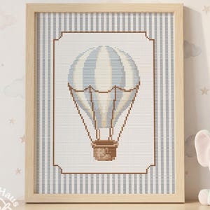 May include: A framed cross-stitch artwork depicting a blue and white hot air balloon. The design is set against a striped background and framed in light wood. A plush elephant toy is placed beside it.
