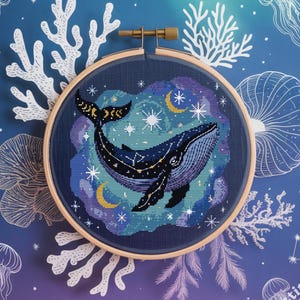 Puede incluir: Un bastidor de bordado de punto de cruz con un diseño de ballena. La ballena es azul oscuro con detalles dorados, sobre un cielo estrellado con lunas crecientes. El fondo es un degradado de azul y morado, con ilustraciones de coral y medusas blancas.